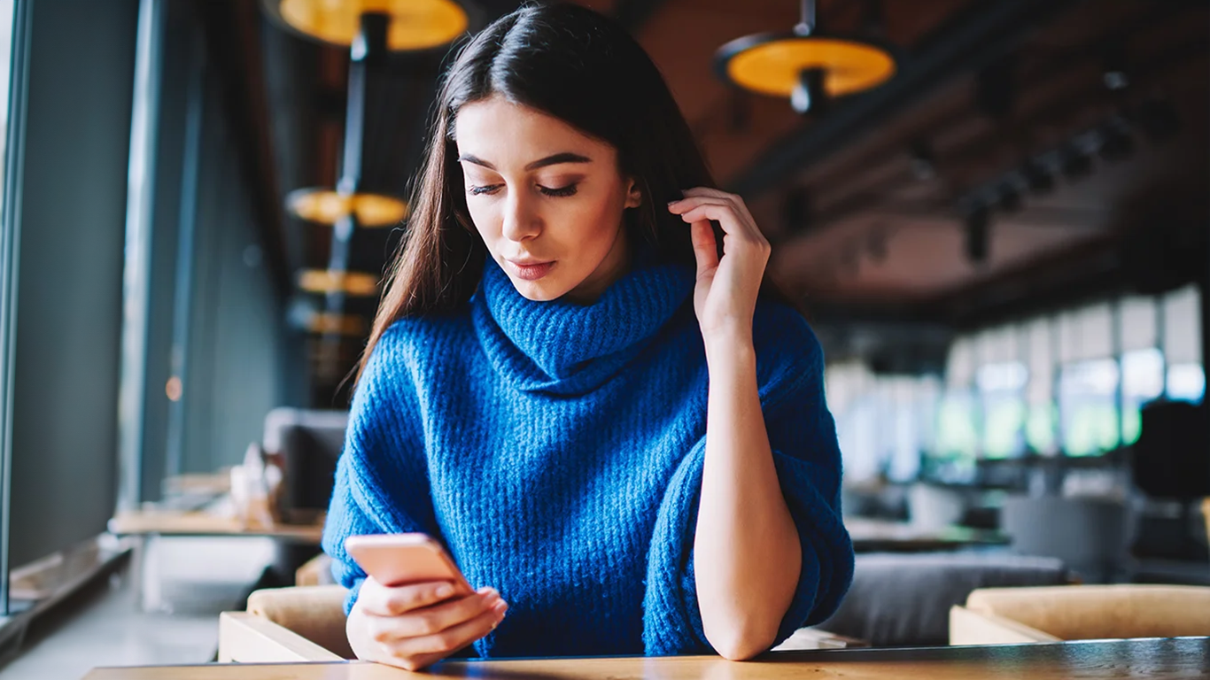 A woman wearing a blue sweater in a modern looking office setting checks her phone